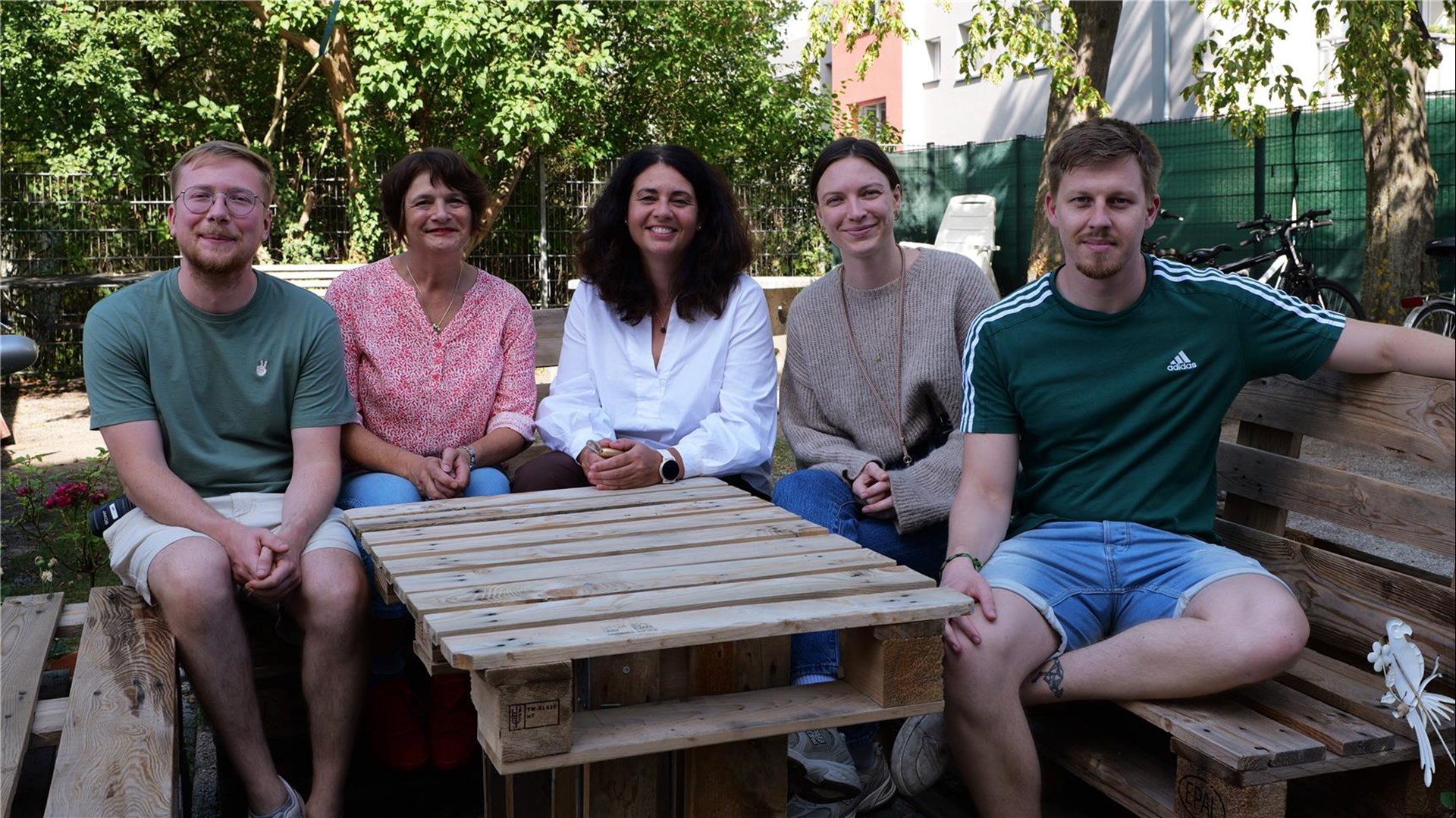 Gruppenfoto von mehreren Menschen, die um einen Holztisch sitzen und in Richtung Kamera schauen.